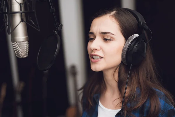 The girl in the recording studio sings a song. Her headphones are on her head. Next to her is a microphone. She emotionally sings the song.