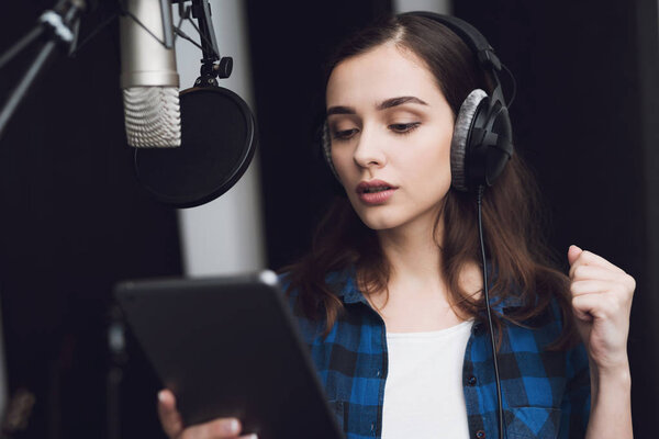 The girl in the recording studio sings a song. Her headphones are on her head. Next to her is a microphone. She emotionally sings the song.