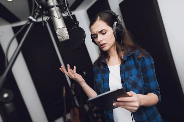 The girl in the recording studio sings a song. She has headphones on her head, and a tablet with text in her hands. Next to her is a microphone. She emotionally sings the song.