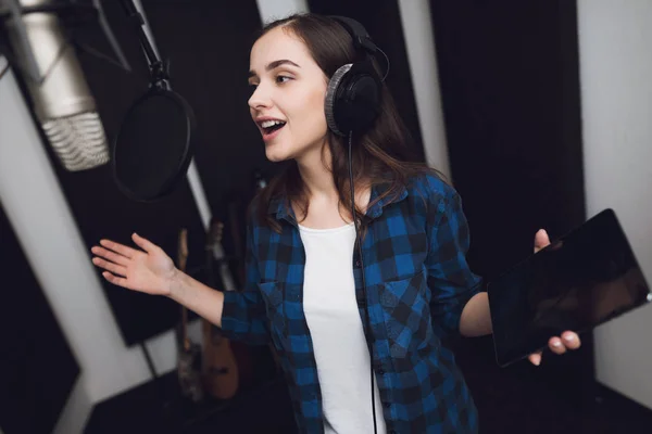 The girl in the recording studio sings a song. She has headphones on her head, and a tablet with text in her hands. Next to her is a microphone. She emotionally sings the song.