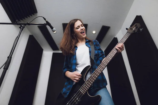 The girl is recording a song at a modern recording studio. She sings to the guitar. There is a microphone in front of her, she has an electric guitar in her hands.