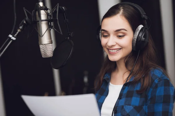 The girl in the recording studio sings a song. Her headphones are on her head. Next to her is a microphone. She emotionally sings the song.