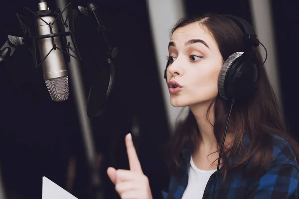 The girl in the recording studio sings a song. Her headphones are on her head. Next to her is a microphone. She emotionally sings the song.