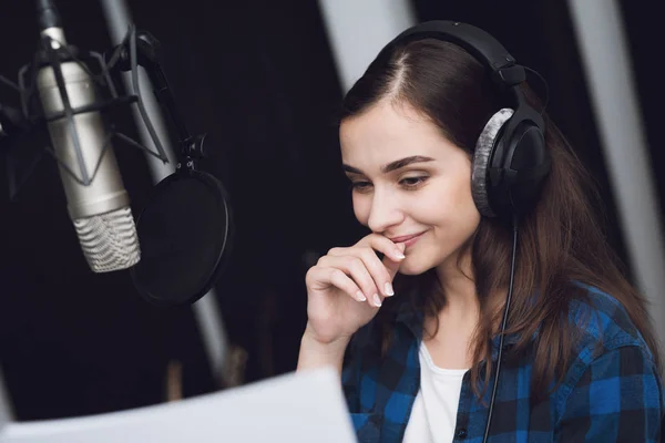 The girl in the recording studio sings a song. Her headphones are on her head. Next to her is a microphone. She emotionally sings the song.