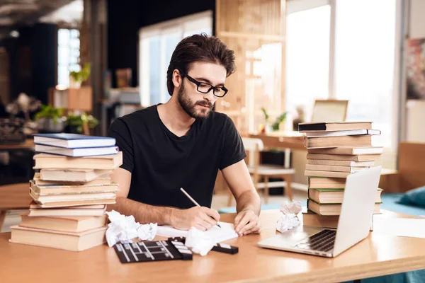 Freelancer γενειοφόρος άνδρας λήψη σημειώσεων που κάθεται στο γραφείο περιβάλλεται από βιβλία. — Φωτογραφία Αρχείου