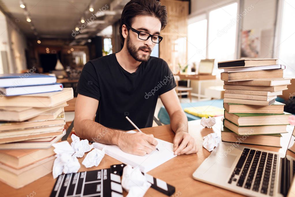 Freelancer bearded man taking notes sitting at desk surrounded by books.