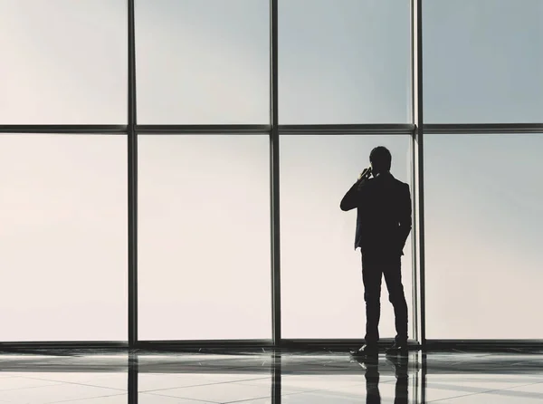 Silhouette back view of young businessman is looking out of a panoramic window and talking by phone — Stock Photo, Image