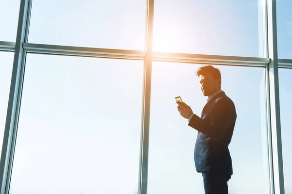 Side view of young businessman is standing near a panoramic window and looking at the phone — Stock Photo, Image