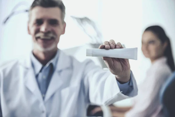 Porträtt av en leende övertandläkare holding tandkräm på bakgrunden hans patient i tandvårdsklinik — Stockfoto