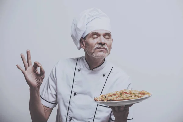 Chefe do sexo masculino cozinheiro em uniforme gestos ok sinal e segurando pizza no fundo cinza — Fotografia de Stock