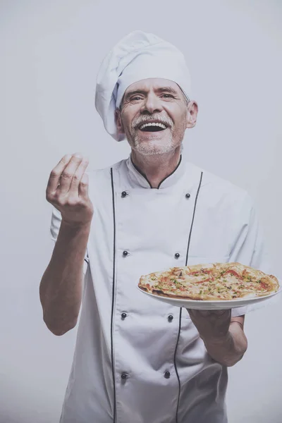 Starší muž šéfkuchař v uniformě drží pizzu na šedém pozadí — Stock fotografie