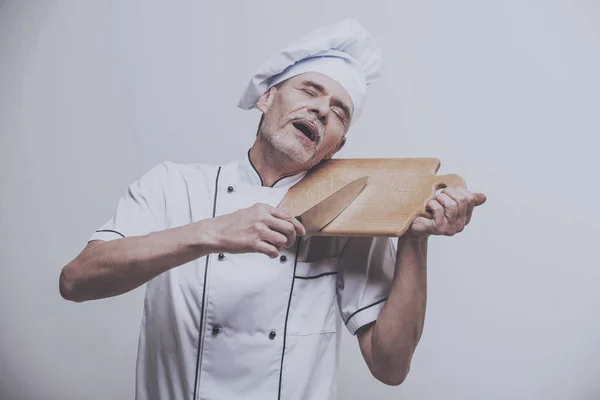 Senior mannelijke chef-kok in uniforme bedrijf mes en hakken aan boord van een op grijze achtergrond — Stockfoto