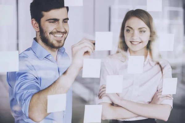 Young businessman and businesswoman working on new project with many sticky notes on glass screen in office — Stock Photo, Image