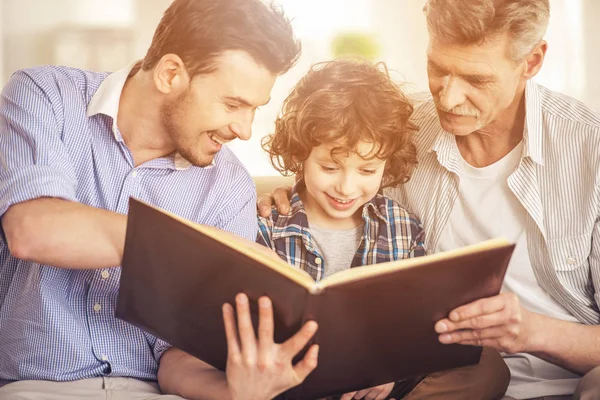 Retrato de generación. Abuelo, padre e hijo sentados y leyendo un libro en el sofá — Foto de Stock