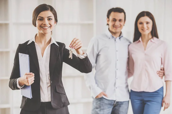 Makelaar vrouw houdt sleutels en jonge koppel op achtergrond — Stockfoto