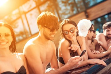 Company of young friends look at photos taken on smartphone in pool in the summertime. Company of young people spend weekend in pool.