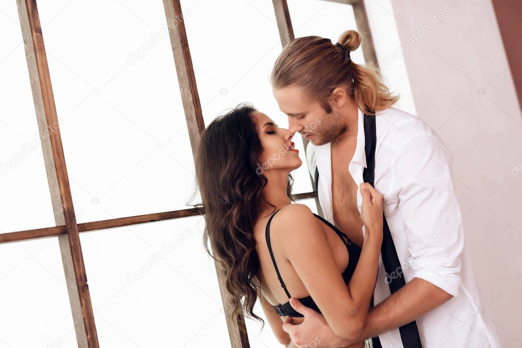Longhaired man gently hugs beautiful woman in lingerie, standing near stained-glass window. Preliminary caresses. Intimate affinity.