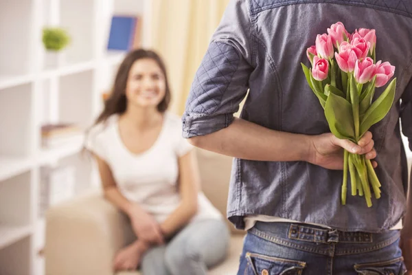 Guy is holding flower behind him is trying to surprise his is waiting girlfriend at home