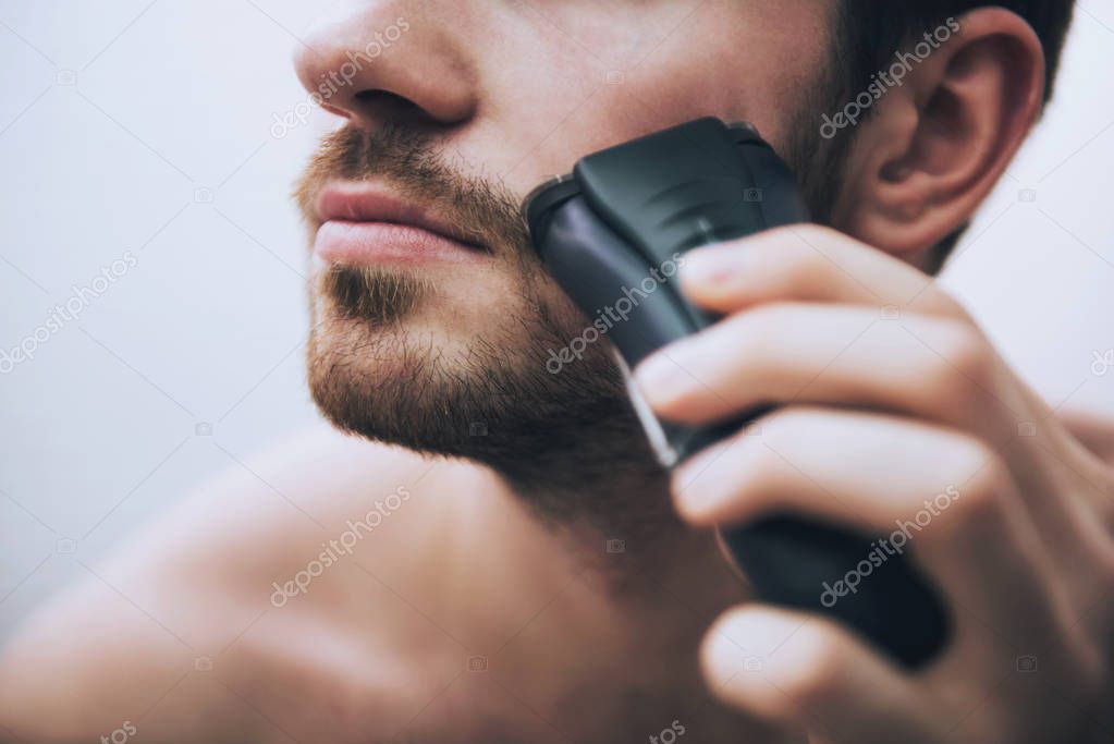 Man is shaving his face with electric razor in bathroom in morning.