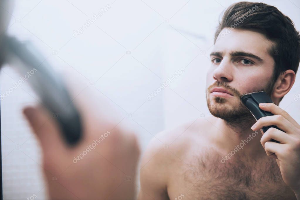 Man is shaving his face with electric razor in bathroom in morning.