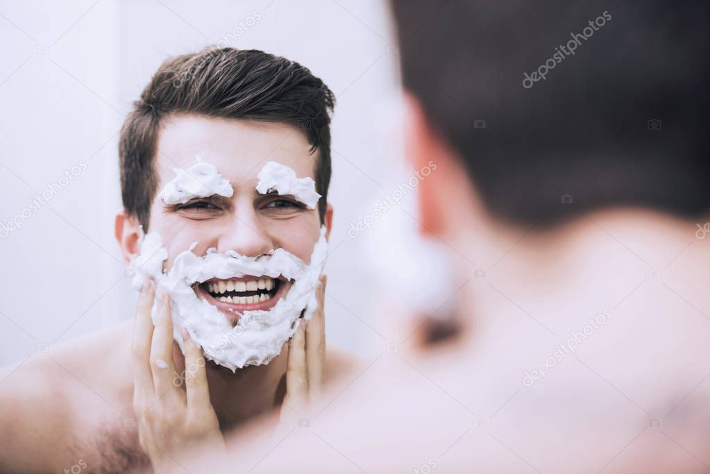 Man is applying shaving foam to his face and brows in bathroom in morning.