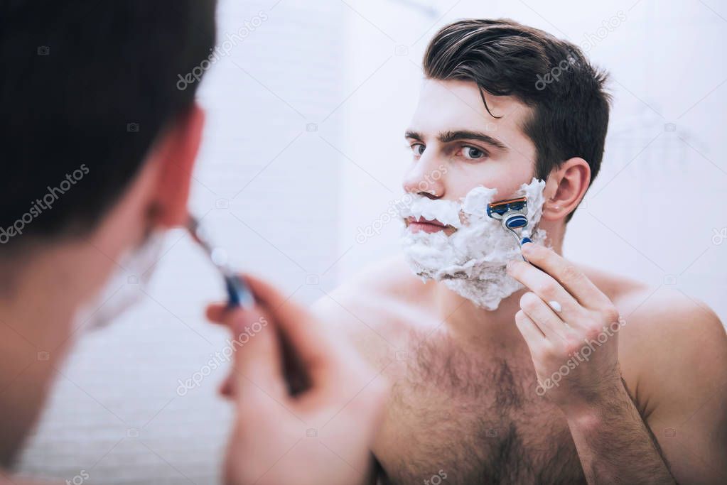 Man is shaving his face looking in mirror in bathroom in morning.
