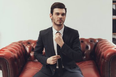 Kendine güvenen genç işadamı kravat izole kırmızı deri koltukta otururken düzeltiyor.