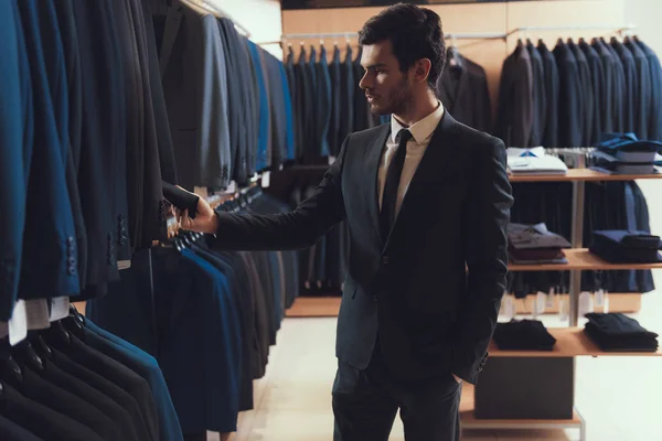 L'homme confiant choisit la veste d'affaires parmi les vêtements suspendus aux cintres dans le magasin de vêtements . — Photo
