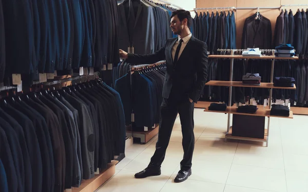 Уверенный человек выбирает деловую куртку среди одежды, висящей на вешалках в магазине одежды . — стоковое фото