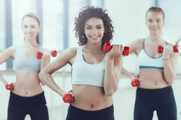 Multinational company of women swinging biceps with dumbbells in gym.