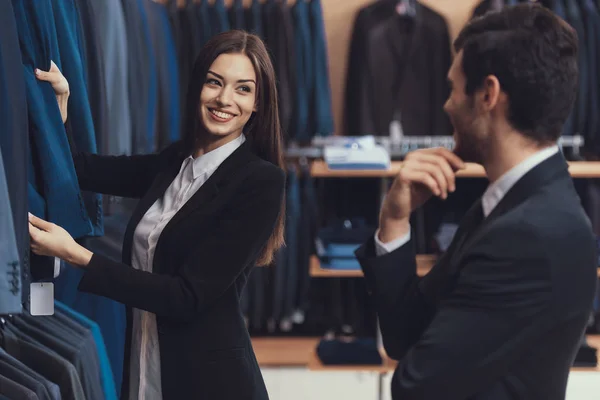 Hermosa mujer consultor de la tienda de ropa masculina ayuda a elegir la chaqueta para el hombre joven en traje . — Foto de Stock