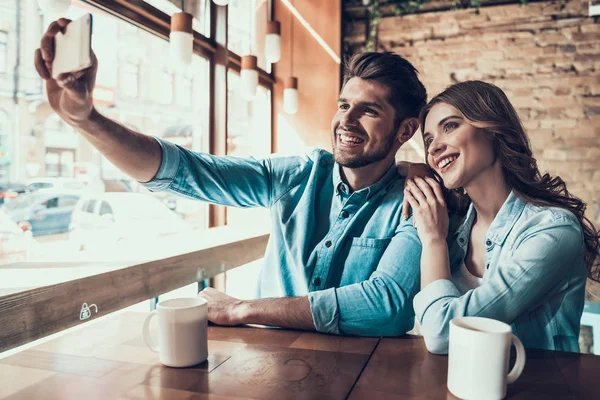 Joven pareja moderna tomando fotos de sí mismos en el teléfono, sentado en la mesa en la cafetería . — Foto de Stock