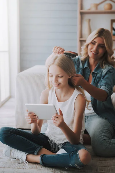 Зріла блондинка мати розчісує миле волосся дочки, яке дивиться на планшет . — стокове фото