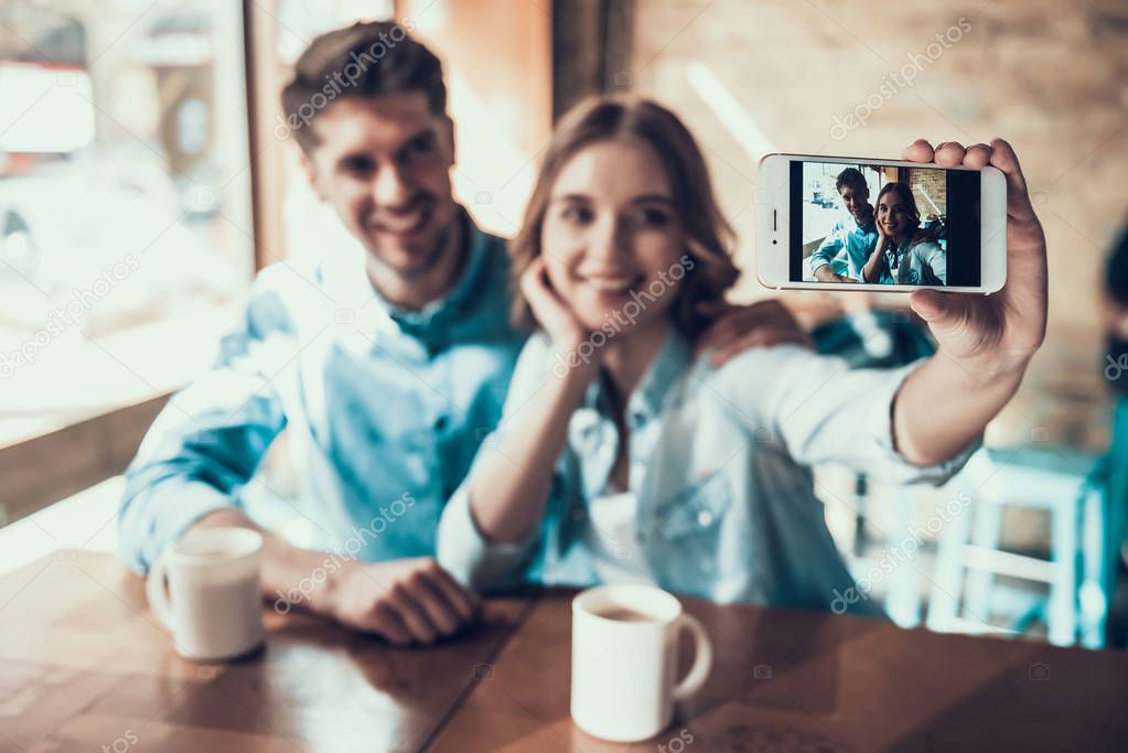 Young modern couple taking pictures of themselves on phone, sitting at table in cafe.