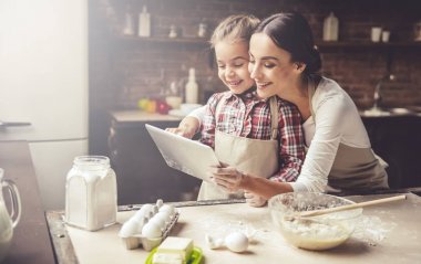 Güzel genç anne ve sevimli küçük kızı dijital tablet kullanma ve evde mutfak pişirme sırasında gülümseyen