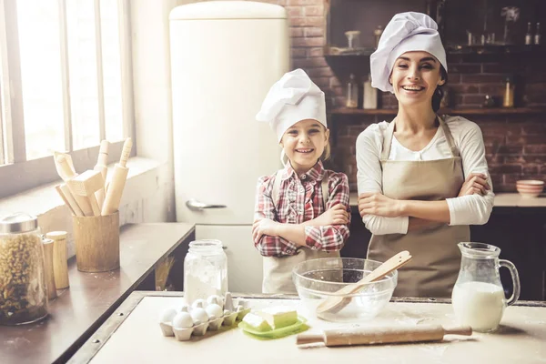 Sevimli küçük kız ve güzel annesi chef şapka içinde çapraz kolları ile ayakta, kameraya bakıyor ve evde pişirme sırasında gülümseyen