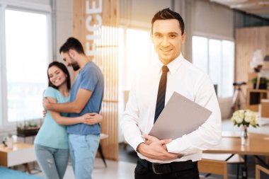 Bir kravat bir Yöneticisi kameraya poz veriyor. Yanına bir adam ve bir kız kucaklayan vardır.