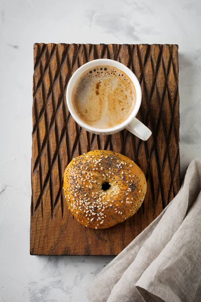 Bagel and a cup of coffee for breakfast on a wooden stand on a light stone or concrete background. — Stock Photo, Image