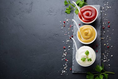 Üç sos seti - mayonez, hardal ve ketçap beyaz seramik kaselerde siyah taş veya beton arka planda. Seçici odaklanma. Üst görünüm.