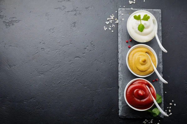 Üç sos seti - mayonez, hardal ve ketçap beyaz seramik kaselerde siyah taş veya beton arka planda. Seçici odaklanma. Üst görünüm.