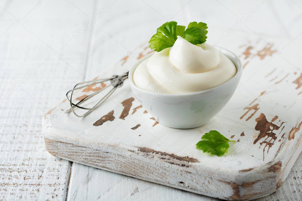 Traditional mayonnaise sauce in white ceramic bowl and ingredients for its preparation on white wooden background. Selective focus.