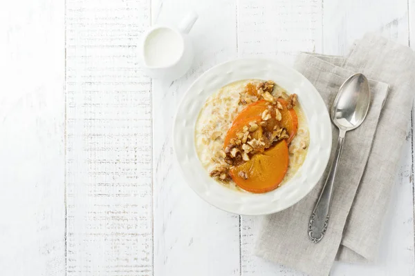 Karamelize Trabzon hurması ve ceviz Beyaz ahşap zemin üzerinde hafif bir seramik kase ile yulaf lapası. Seçici odak. Üstten Görünüm. Kopya alanı.