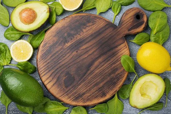 Raw fresh avocado, spinach leaves and lemon with empty wooden cutting board on dark old concrete or stone background. Selective focus. Top view. Copy space.