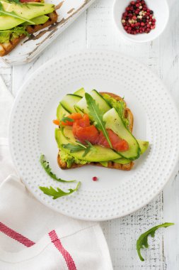 Alabalık, somon, avokado, salatalık ve roka ahşap stand Beyaz ahşap masa arka plan üzerine açık tost. Seçici odak. Üstten Görünüm. Kopya alanı