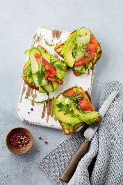 Alabalık, somon, avokado, salatalık ve roka ahşap stand gri beton zemin üzerine açık tost. Seçici odak. Üstten Görünüm. Kopya alanı