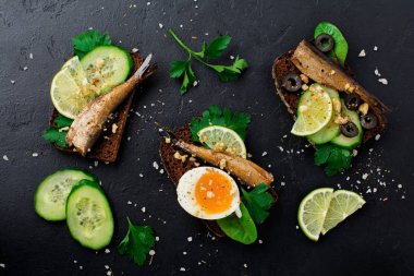 Sandviç hamsi, salatalık, kireç, haşlanmış yumurta, maydanoz yaprakları ve pazı siyah eski beton veya taş arka plan üzerinde siyah ekmek üzerinde balık. Seçici odak. Rustik tarzı. Üstten Görünüm. Kopya alanı.