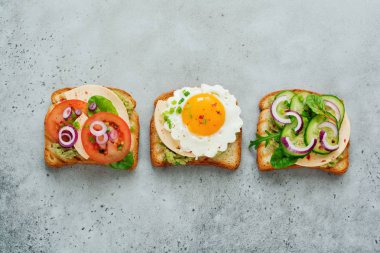 Üç farklı guacamole sos, sahanda yumurta, taze salatalık, peynir ve domates pazı, roka, gri beton zemin üzerine kırmızı soğan ile sandviçler tost. Seçici odak.