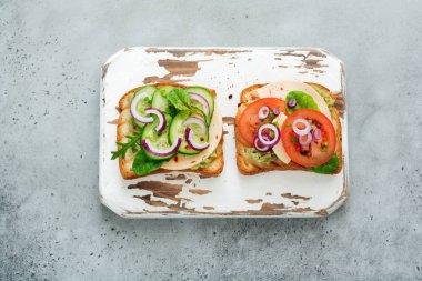 Guacamole sos, taze salatalık, peynir ve domates pazı, roka, gri beton zemin üzerine kırmızı soğan ile iki farklı tost sandviç. Seçici odak.