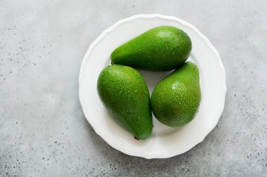 Gri beton zemin üzerine beyaz eski seramik tabak içinde üç ham taze avokado. Seçici odak. Üstten Görünüm.