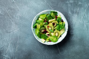 Ispanak bebek salata yaprakları, su teresi, kivi, avokado ve yaşlı seramik plaka gri beton zemin üzerine nar. Seçici odak. Üstten Görünüm. Kopya alanı.